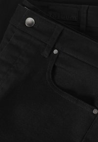 Gros plan sur un jean en denim noir montrant la poche avant, les rivets en argent, les passants de ceinture et les détails de la fermeture par bouton.