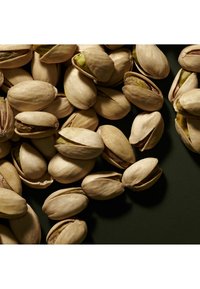 Orzechy pistacjowe w beżowych łupinach, niektóre otwarte, ukazujące zielone jądra, rozrzucone na ciemnozielonej powierzchni. Gładka tekstura z widocznymi szwami.