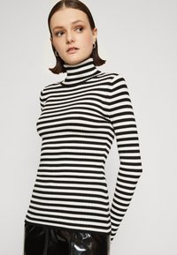 Črno-bela prugasta turtleneck pulover s rebrasto teksturo, prilagojenim krojem in dolgimi rokavi, v kombinaciji s sijočimi črnimi hlačami.