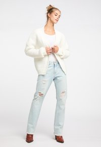 Weißer gestrickter Cardigan mit strukturiertem Muster, kombiniert mit hellblauen, ausgewaschenen Jeans und braunen Absatzstiefeletten.