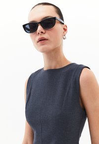 Gafas de sol en forma de ojo de gato negras con montura ancha, lentes oscuros y detalles plateados. Llevadas con un vestido sin mangas de color gris oscuro. Textura suave.