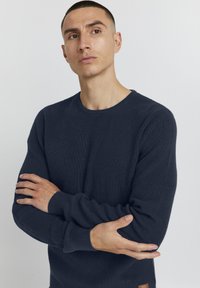 Pull en maille bleu marine avec une texture côtelée, un col rond et des manches longues. Présente une petite étiquette de marque près de l'ourlet.