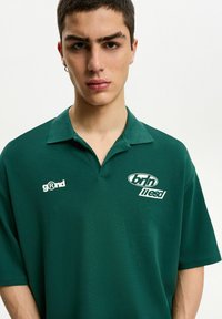 Mörkgrön pikétröja med korta ärmar, krage och vit logotyp på bröstet. Mjuk tyg med avslappnad passform.