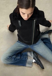 Czarna kurtka z owczej wełny z zamkiem błyskawicznym, z szerokim kołnierzem i skórzanymi akcentami, w połączeniu z rozszerzanymi jasnoniebieskimi dżinsami i czarnymi mokasynami.