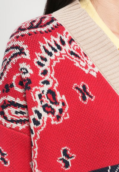 Pull en tricot rouge avec un design texturé et des motifs noirs et crème. Le col beige côtelé ajoute un contraste à la couleur vive.