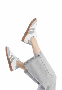 Zapatillas blancas con detalles en gris, de material de cuero, con punta redonda y suela de goma texturizada. Combinadas con unos jeans anchos grises.