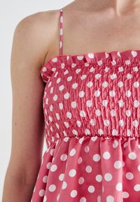 Pink sundress featuring white polka dots, smocked bodice, and ruffled neckline. Thin shoulder straps and flared skirt detail.
