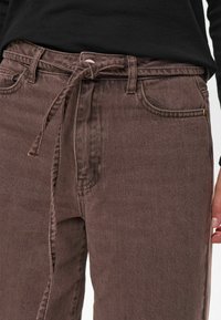 Rjave denim hlače s prednjim trakom za zavezovanje, klasičen pet-pocket design in teksturirana površina. Vidne podrobnosti šivanja ob šivih.