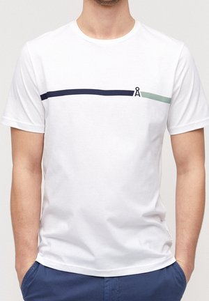 Weißes T-Shirt mit kurzen Ärmeln und horizontalen Linien in Marineblau und Hellgrün, getrennt durch ein kleines schwarzes Kreissymbol, getragen von einer Person mit in die Taschen gesteckten Händen.