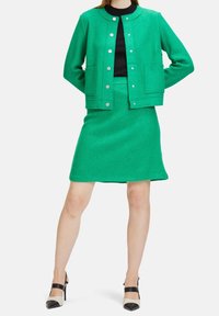 Grön texturerad jacka med silverknappar och två framfickor, matchad med en grön kjol. Modellen bär en svart topp och tvåtonade klackskor.