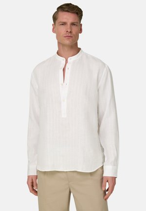 Mand iført en hvid langærmet hørskjorte med en bandekrave og beige bukser, stående mod en ensfarvet baggrund.