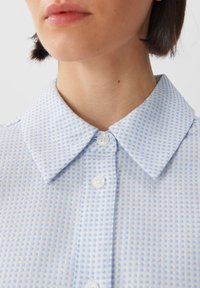 Ljusblå mönstrad knäppskjorta med en texturerad rutdesign, med spetsig krage och vita knappar. Närbild av halsringningen.