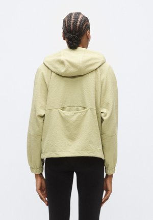 Veste vert clair texturée avec capuche, épaules tombantes, poignets élastiques et grande poche arrière. Design simple, accessoires minimaux.