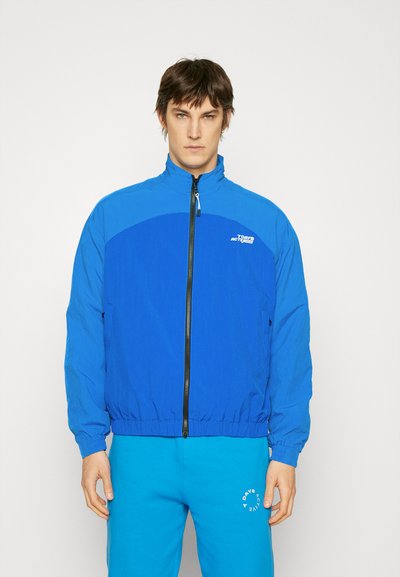 Kék széldzseki magas gallérral, teljes cipzárral, rugalmas mandzsettákkal és derékkal, sötétebb kék panelek és logó a mellkason.