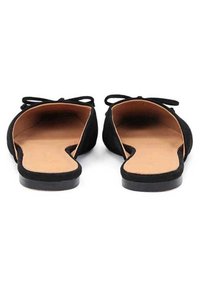 Black suede flats with a rounded toe, open sides, and a bow accent on the heel. Brown leather insole and a flat black sole.