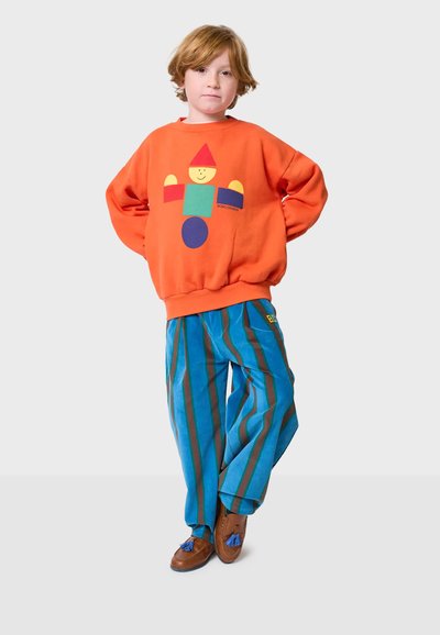 Sudadera naranja con estampado geométrico juguetón; pantalones a rayas azules de tela suave; zapatos marrones adornados con borlas.