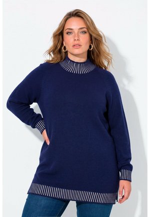 Kvinde med lyst brunt hår iført en mørkeblå sweater med hvide striber på kraven, manchetterne og kanten, poserer med den ene hånd på hoften.