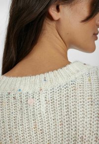 Strickpullover mit lockerem, strukturiertem Gewebe in Creme, das farbige Sprenkel und ein pinkes Knopfdetail am rückseitigen Halsausschnitt aufweist.