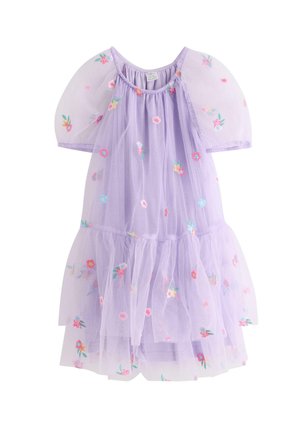 Ung pige smiler, iført en lys lilla kjole med puffede ærmer og farverig blomsterbroderi, stående på en ensfarvet hvid baggrund.