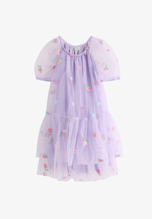 Giovane ragazza sorridente, che indossa un vestito viola chiaro con maniche a sbuffo e ricami floreali colorati, in piedi su uno sfondo bianco uniforme.