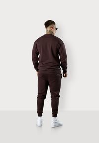 Braunes Sweatshirt- und Jogginghosen-Set, hergestellt aus weichem Stoff. Gerippte Bündchen und Taillengummi, kombiniert mit weißen Sneakers. Dezentes Logo-Detail auf der Hose.