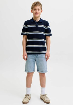 Ragazzo in piedi, indossa una polo a righe blu navy, pantaloncini di jeans chiari, calzini bianchi e scarpe beige con lacci, sorride alla fotocamera.