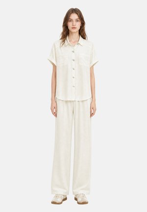 Jeune femme debout portant une chemise blanche ample à manches courtes boutonnée et un pantalon large avec des baskets blanches et beiges.