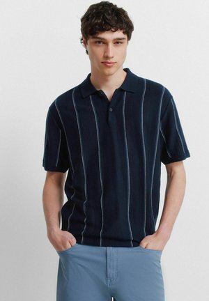 Junger Mann trägt ein marineblaues kurzärmeliges Poloshirt mit vertikalen Streifen und hellblauen Hosen, Hände in den Taschen, steht vor einem weißen Hintergrund.