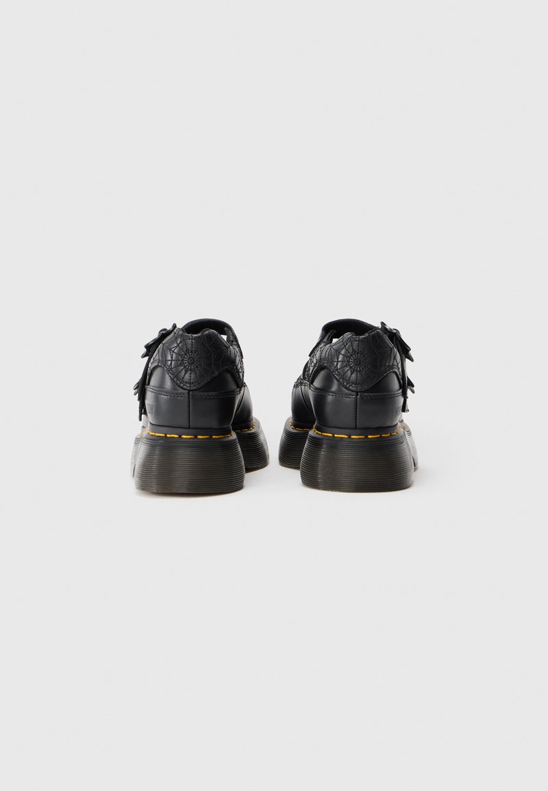 Une paire de chaussures en cuir noir chunky avec des semelles épaisses et des coutures jaunes, vue de l'arrière sur un fond blanc uni.