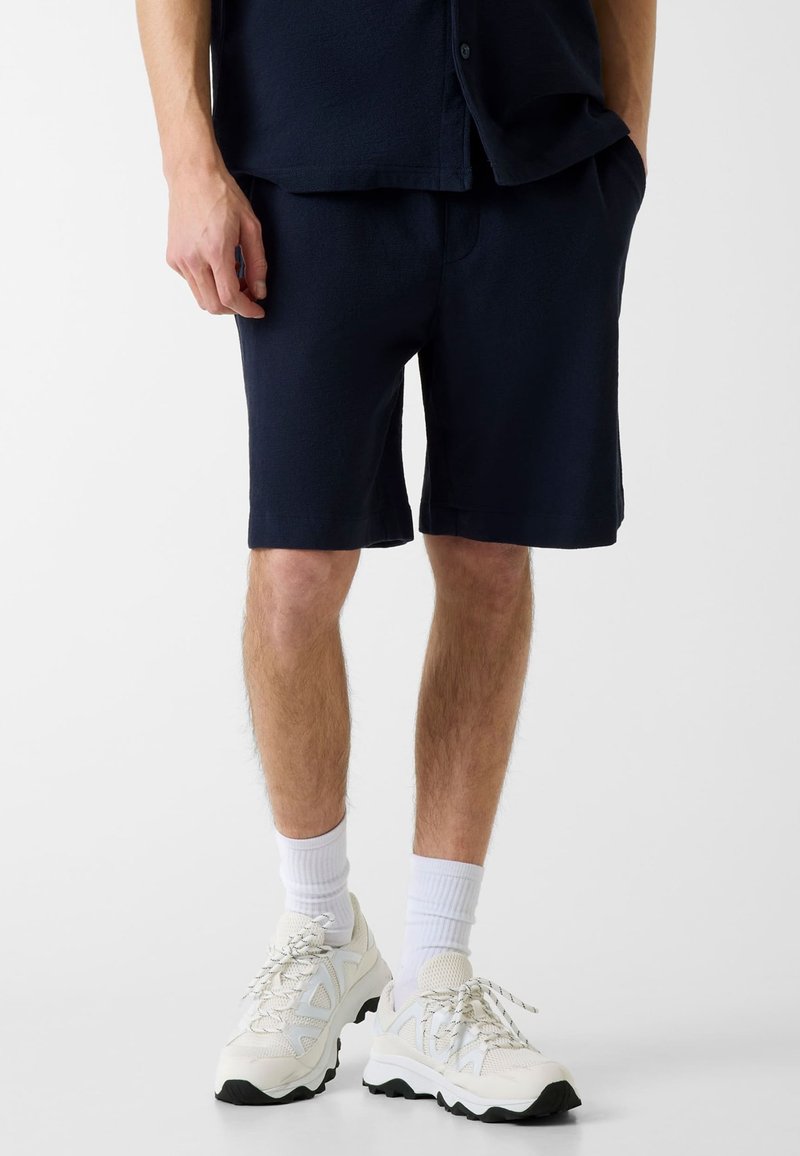 Personne portant un short bleu marine, des chaussettes blanches à mi-mollet et des baskets blanches volumineuses, debout devant un fond blanc uni.