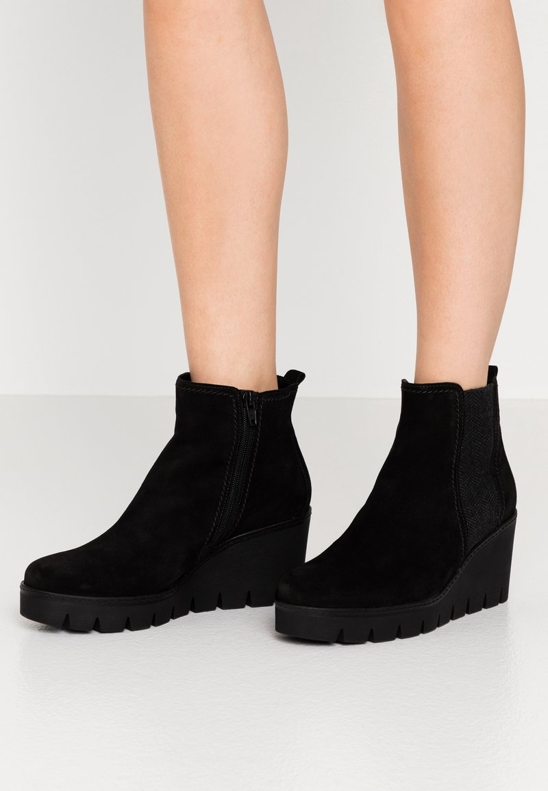 Bottines noires en daim compensées avec fermeture éclair latérale et panneau élastique texturé, portées sur jambes nues sur fond uni.