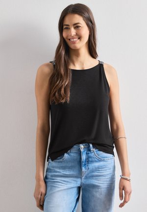 Femme souriante aux longs cheveux bruns, portant un haut noir sans manches avec des bretelles rayées et un jean bleu clair, debout contre un mur uni.