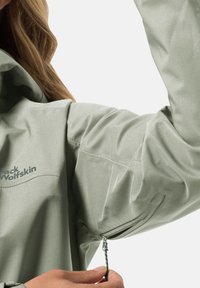 Giacca impermeabile verde chiaro con una texture morbida, polsini regolabili con zip e dettaglio del logo sul lato sinistro anteriore.