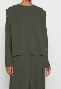 Mørkegrønt oversized top med lange ærmer, rund hals og skulderplisseringer. Stoffet ser glat ud med en subtil glans. Accentknapdetalje.
