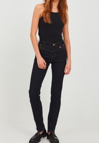 Dark blue denim jeans with a fitted shape, five pockets, and contrasting orange stitching. Worn with a black tank top and black shoes.