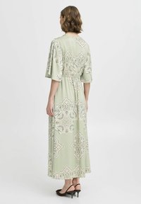Rochie maxi din material verde deschis cu model paisley, cu mâneci scurte, talie elastică și design fluid. Asortată cu sandale negre cu toc.