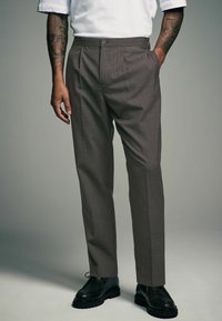 Brown pinstriped trousers featuring a straight fit, front pleats, side pockets, and a button closure, paired with black lace-up shoes.