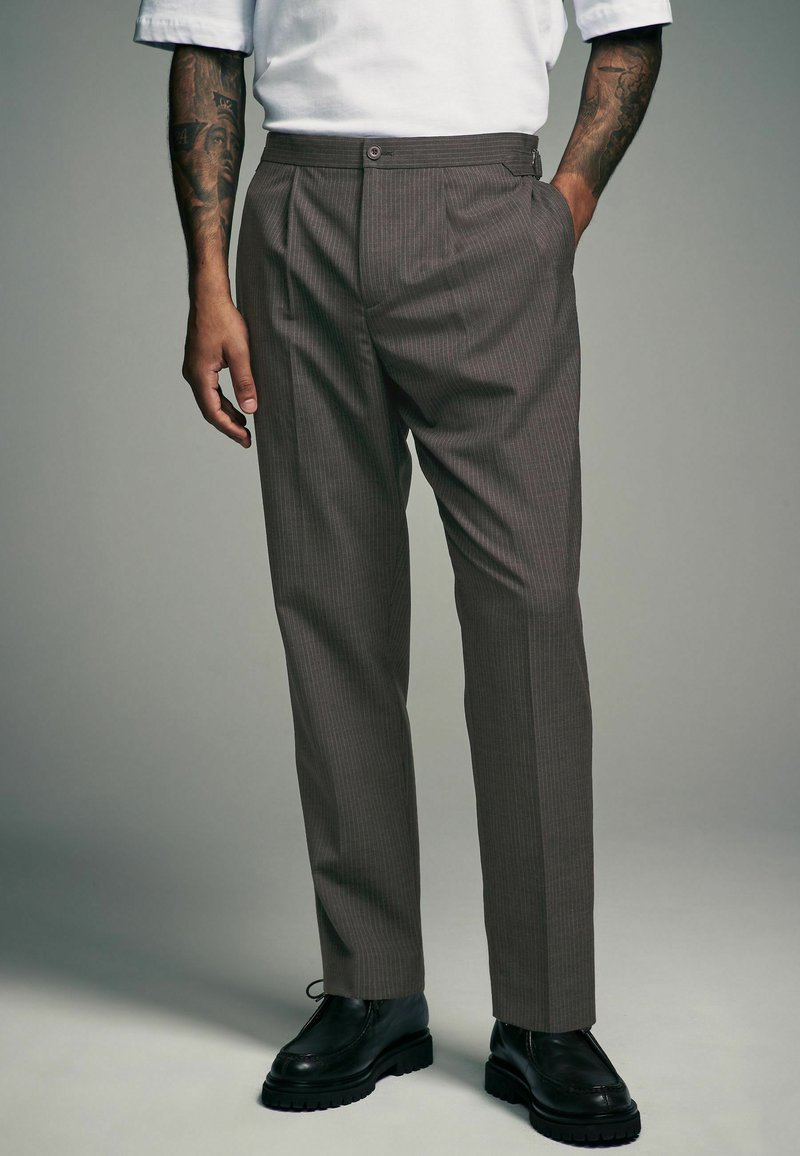 Pantalons marron à rayures fines avec une coupe droite, des plis sur le devant, des poches latérales et une fermeture à bouton, associés à des chaussures noires à lacets.