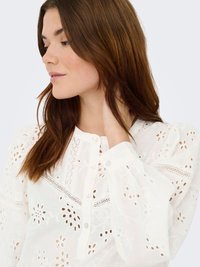 Blouse blanche en matière légère, ornée de broderies florales, fermeture boutonnée, manches longues et fluides avec des détails délicats.