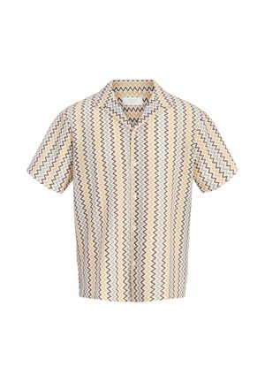Blond chłopiec uśmiechający się, ubrany w koszulę z krótkim rękawem w zygzakowaty wzór na białym t-shircie, ciemne dżinsowe szorty, białe skarpetki i brązowe skórzane buty.
