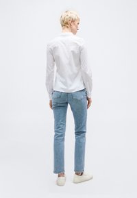 Άτομο με κοντά ξανθά μαλλιά, ντυμένο με λευκό πουκάμισο με μακριά μανίκια, ανοιχτό μπλε τζιν και λευκά αθλητικά παπούτσια, στέκεται γυρισμένο προς τα πίσω σε λευκό φόντο.