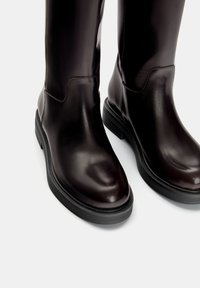 Bottes en cuir marron jusqu'aux genoux avec une texture lisse, un bout rond et une épaisse semelle en caoutchouc noir, mettant en avant un design minimaliste et une fonctionnalité.