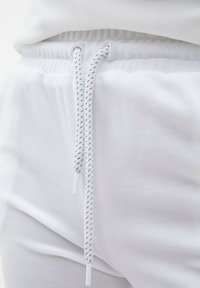 Białe dresowe spodnie z elastycznym pasem, z białym sznurkiem do ściągania o plecionej teksturze i plastikowymi końcówkami, wykonane z miękkiej tkaniny.