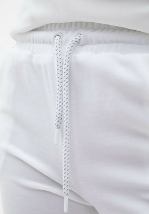 Białe dresowe spodnie z elastycznym pasem, z białym sznurkiem do ściągania o plecionej teksturze i plastikowymi końcówkami, wykonane z miękkiej tkaniny.