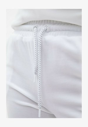 Białe dresowe spodnie z elastycznym pasem, z białym sznurkiem do ściągania o plecionej teksturze i plastikowymi końcówkami, wykonane z miękkiej tkaniny.