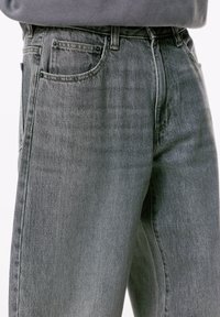 Nahaufnahme einer Person, die hochtaillierte graue Jeans trägt, bei denen die vorderen und Münztaschen sichtbar sind, kombiniert mit einem in die Hose gesteckten grauen Oberteil.