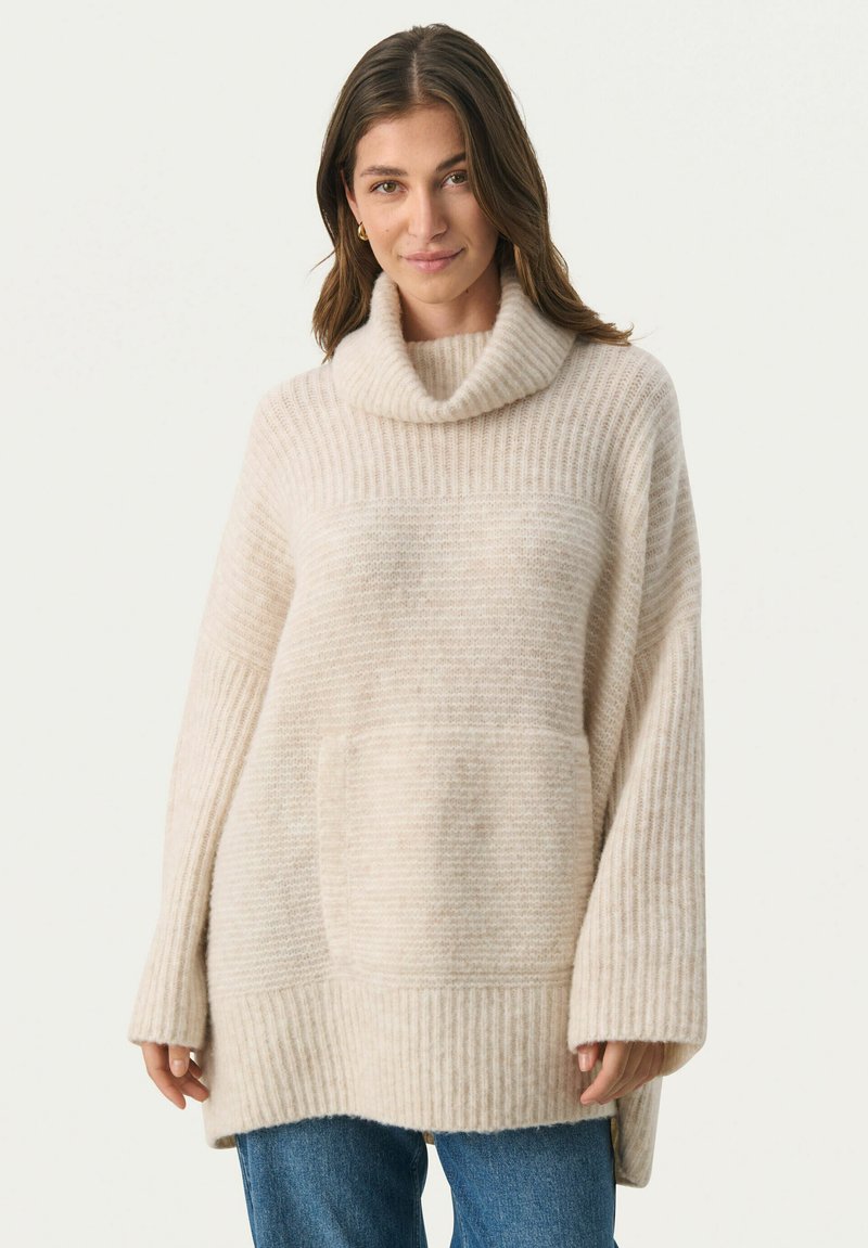 Cremefarvet oversized strikket sweater med høj hals, ribbet tekstur, frontlomme og sænkede skuldre, parret med blå jeans.