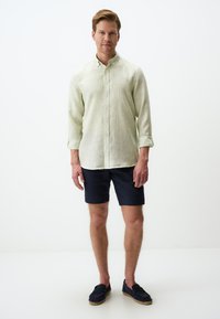Hellgrünes, langärmeliges, knöpfbares Hemd mit strukturiertem Muster; kombiniert mit marineblauen Shorts und dunklen Slippern, stehend vor einem weißen Hintergrund.