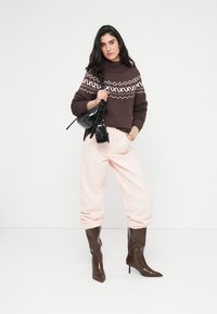 Καφέ πλεκτή ζακέτα με λευκά γεωμετρικά σχέδια, ανοιχτό ροζ φαρδύ παντελόνι και καφέ μπότες με μύτη και τακούνι. Κρατάει μια μικρή μαύρη τσάντα.