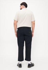 Beige kortærmet polo med krave, sammen med sorte tapered bukser. Sorte sko og hvide sokker synlige. Enkel, afslappet design.